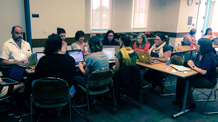 A group of people sitting around tables with laptops in a classroom or workshop setting.