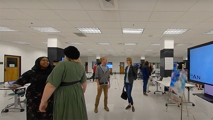 A wide view of a modern exhibition space with people interacting and viewing digital displays. The room is well-lit with white walls and ceilings, featuring large screens, posters on easels, and scattered desks.