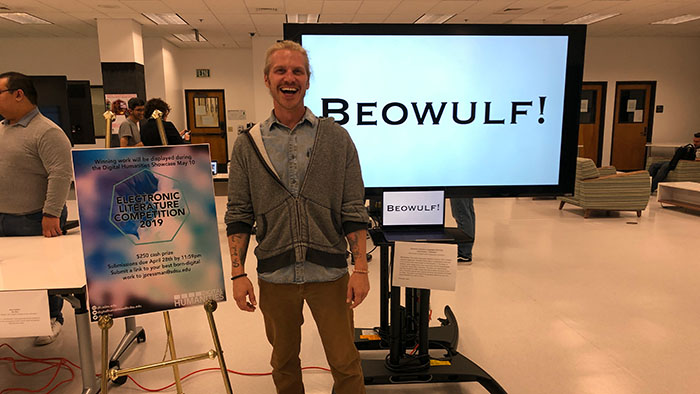 A man stands smiling in front of a screen displaying the word “Beowulf!” with a poster for an Electronic Literature Competition nearby.