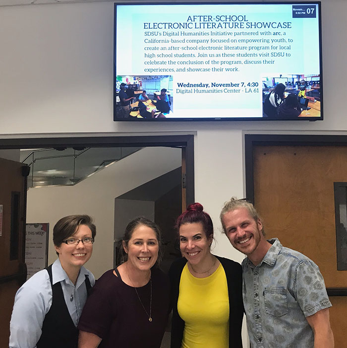 Four smiling people pose in front of a digital screen promoting an “After-School Electronic Literature Showcase” event at SDSU.