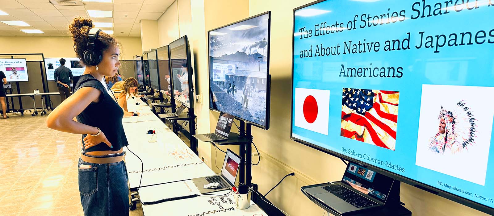 A young woman wearing headphones observes a presentation displayed on a large screen in a brightly lit room. The screen shows a blue slide titled “The Effects of Stories Shared By and About Native and Japanese Americans,” with images of the Japanese flag, the American flag, and a Native American in traditional headdress. Other people are engaged at adjacent screens and workstations in the background.