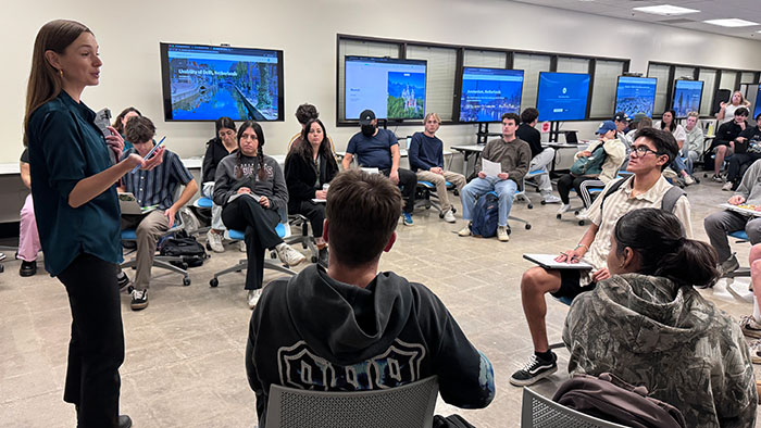 A group of Geography students seated in a circle in the Digital Humanities Center, attentively listening to a professor speaking at the front. There are multiple large screens displaying presentation slides are visible in the background.