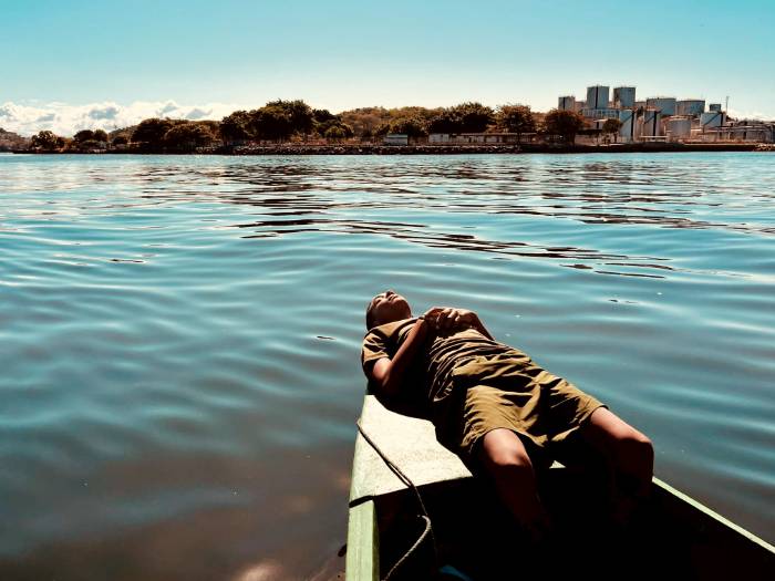 man resting on top of boat in water with city in the background