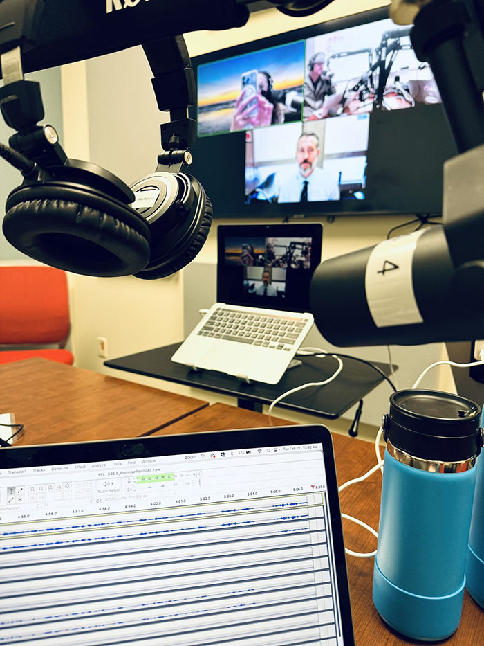 A recording studio setup featuring a laptop with audio editing software, microphones, headphones, and a video call displayed on a large screen.