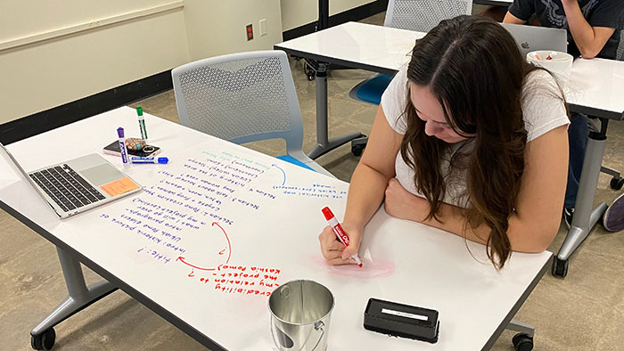 A student writing ideas with a marker on a whiteboard desk, surrounded by notes, a laptop, and supplies.