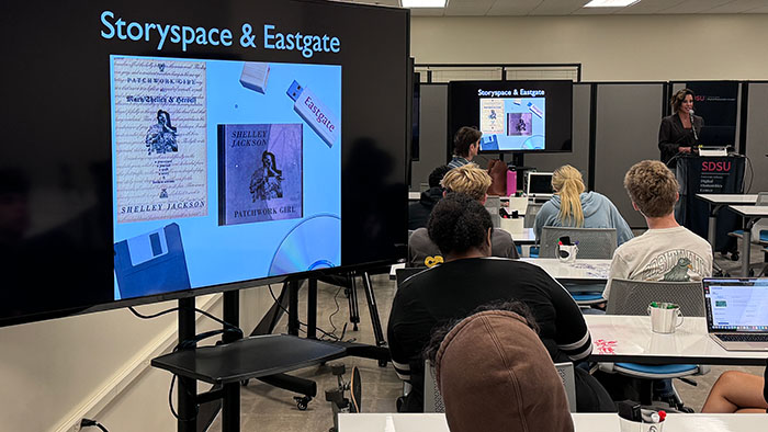 A presenter discussing electronic literature with a large screen showing "Storyspace & Eastgate" to an engaged audience.