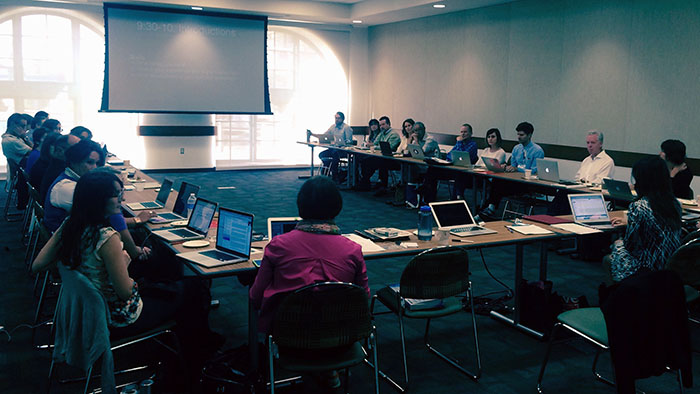 A large group of people sitting around tables in a conference room with laptops and a projector screen.