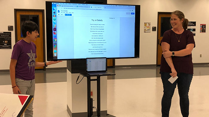 A young person and an adult stand next to a large screen displaying an e-literature poem titled “Try or Satisfy.”