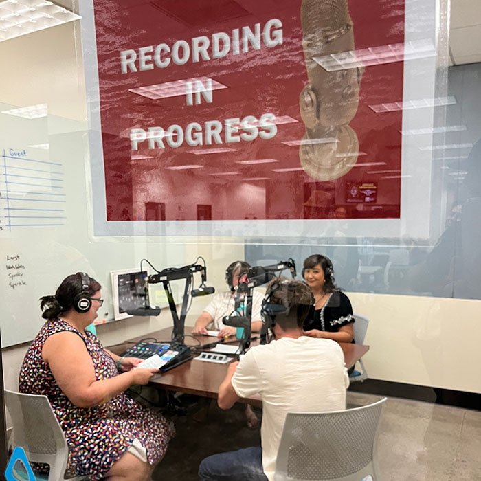 Four people sit at a table in a recording studio with microphones, headphones, and audio equipment under a “Recording in Progress” sign.