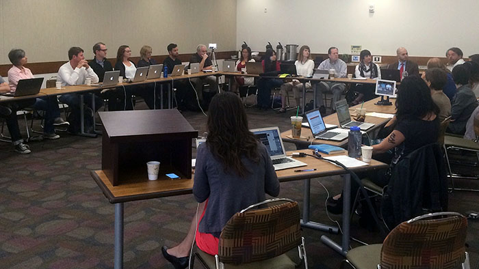 A group of people sitting around tables in a conference room with laptops and papers.