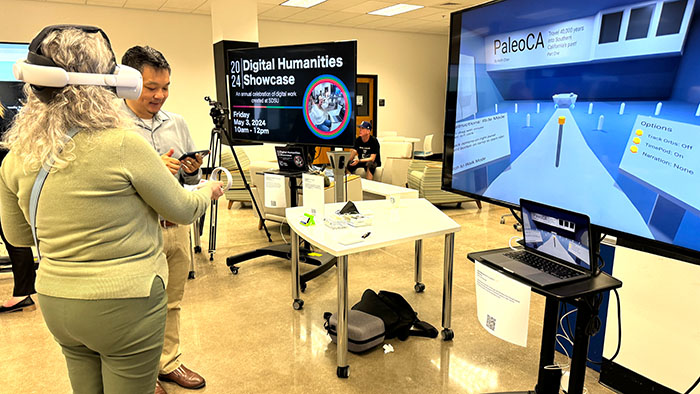 A person uses a VR headset at a Digital Humanities Showcase, with a large screen displaying a virtual interface called “PaleoCA.”