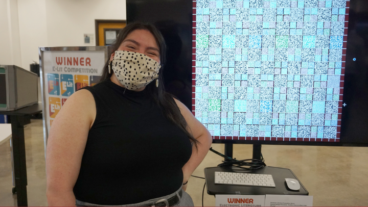 A masked student stands next to a large screen displaying her e-lit project, a digital quilt of QR codes